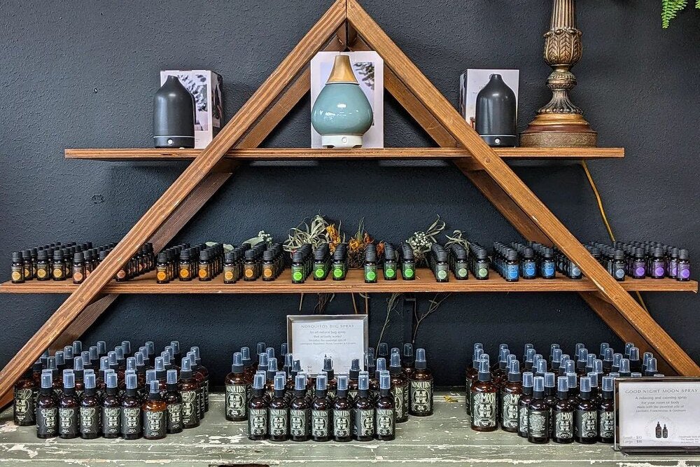 Wooden triangle shelves in The Humble Life holding rows of small amber bottles of essential-oil sprays on a dark wall.
