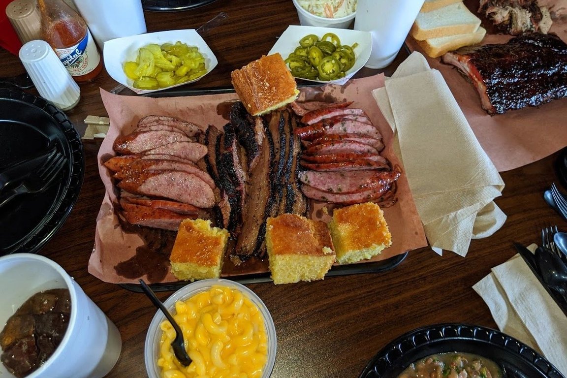 An overhead view of a tray of sliced brisket, sausage and cornbread, with mac and cheese, beans, pickles and jalape&ntilde;os around it