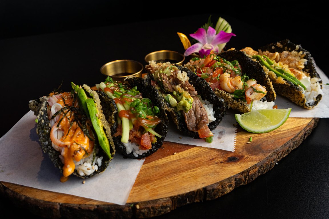 Five crispy nori sushi tacos on a wooden board with sauces, a lime wedge and an orchid garnish