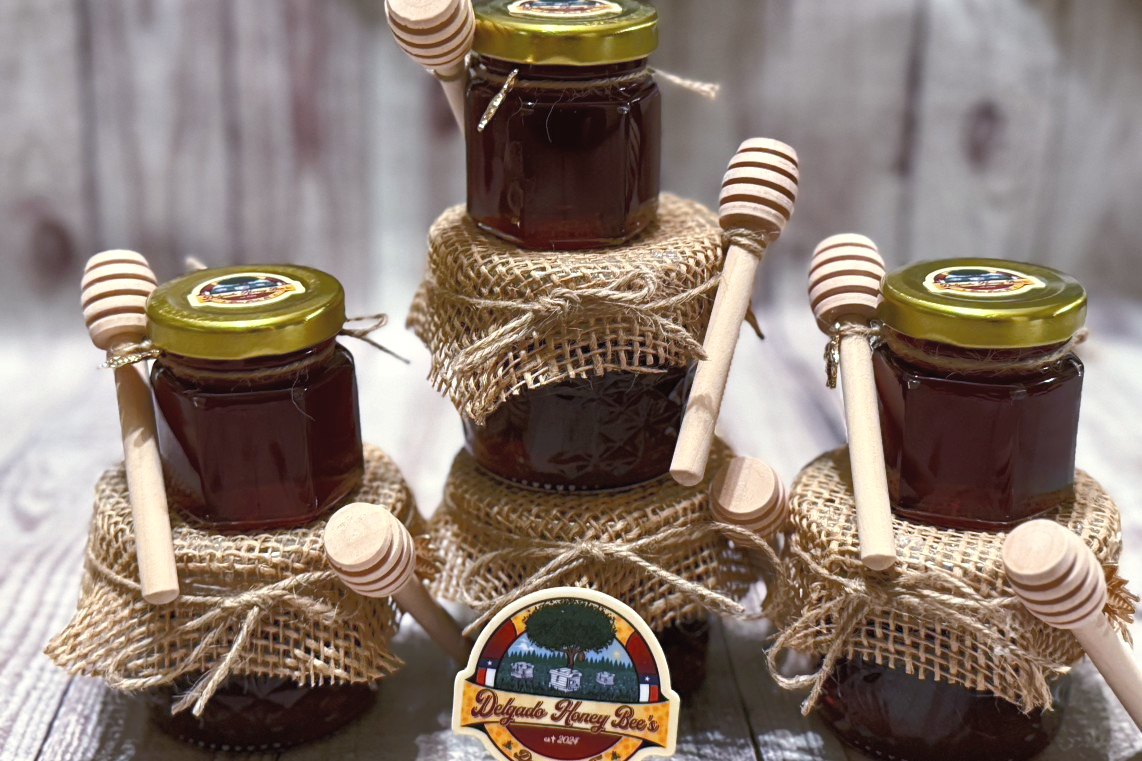 Three small jars of dark honey tied with burlap and wooden dippers, each with a Delgado Honey Bee’s label
