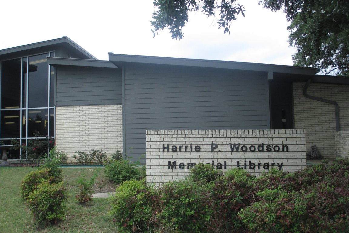 A modern gray library building with a brick sign reading Harrie P. Woodson Memorial Library in front of shrubs.