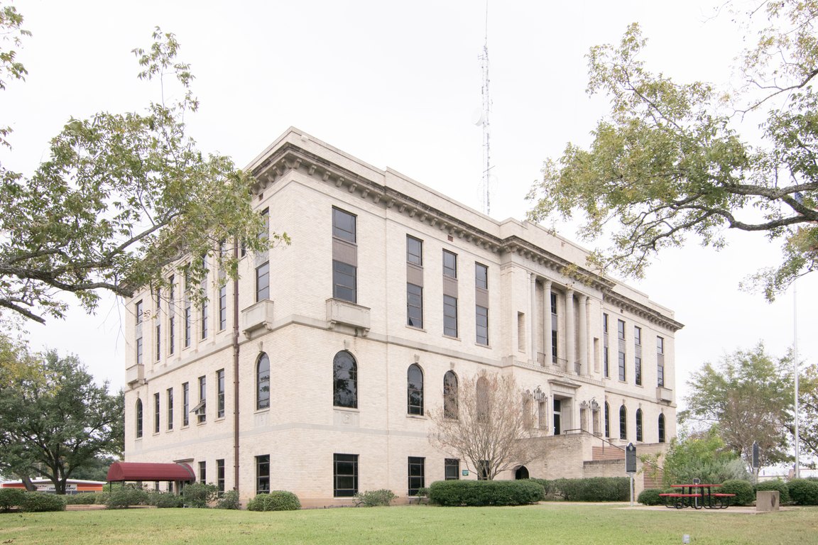 A three-story pale stone courthouse with tall columns and arched windows stands behind live oak trees and a lawn.
