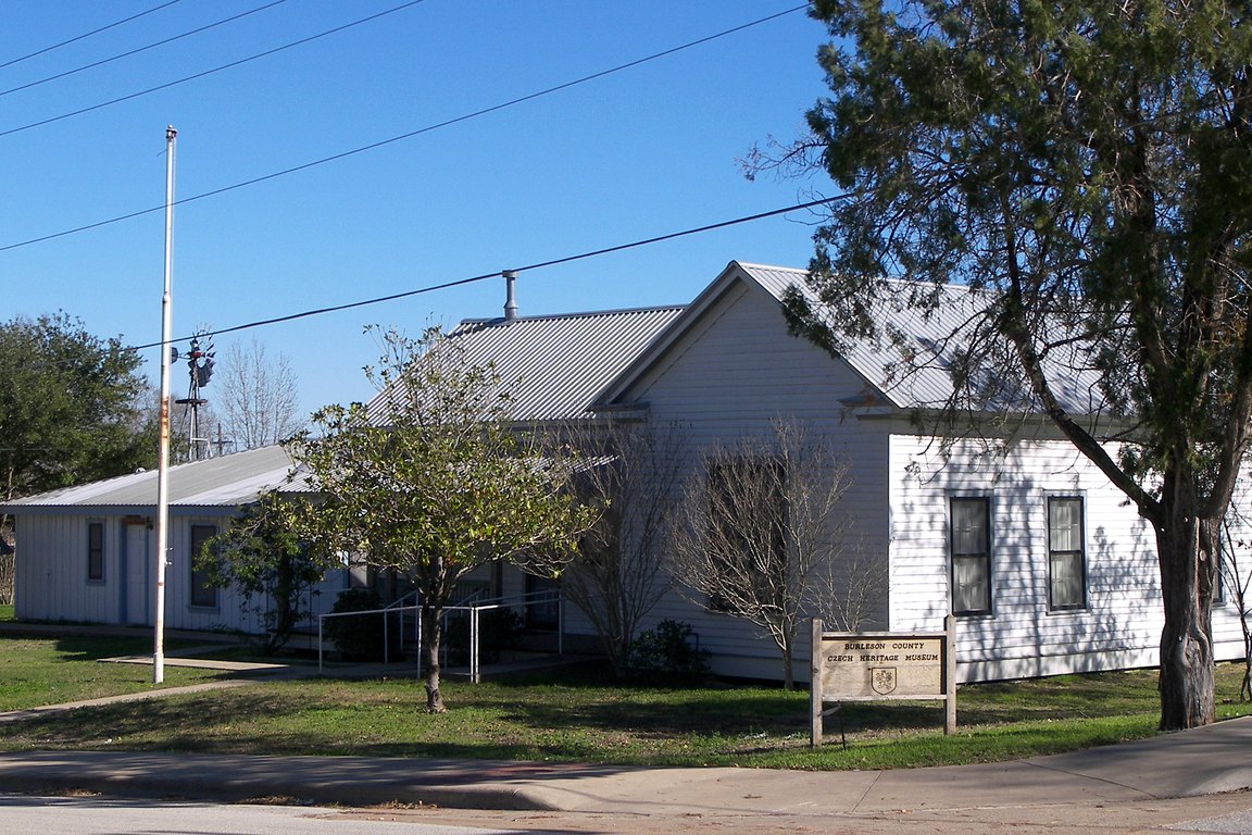 A white wood-frame building with a metal roof and a small sign out front, behind a lawn and trees.