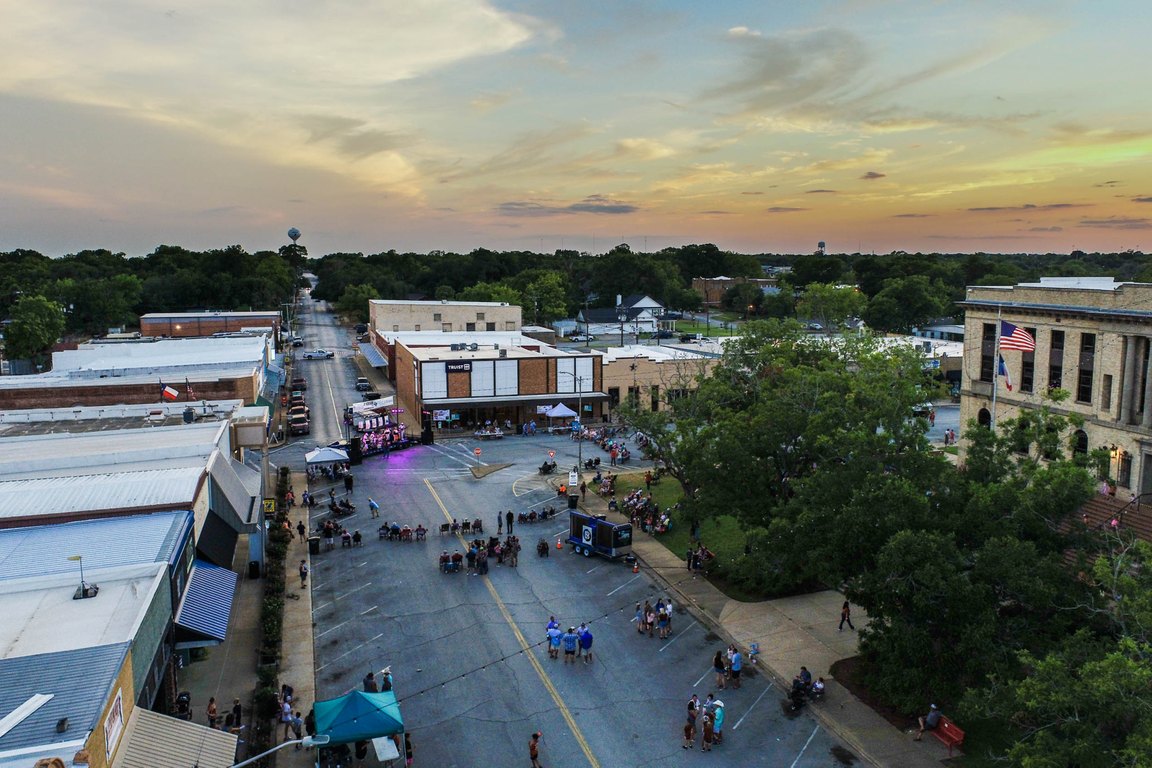An aerial view at dusk of a downtown street lined with low commercial buildings, small crowds and tents, with the courthouse at right.