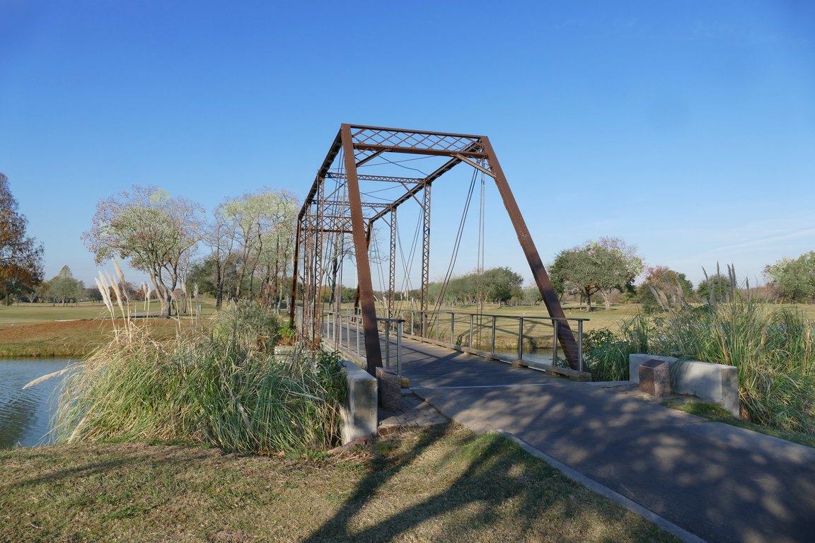 A rust-colored iron truss bridge crosses a pond, with tall grass in the foreground and trees behind.
