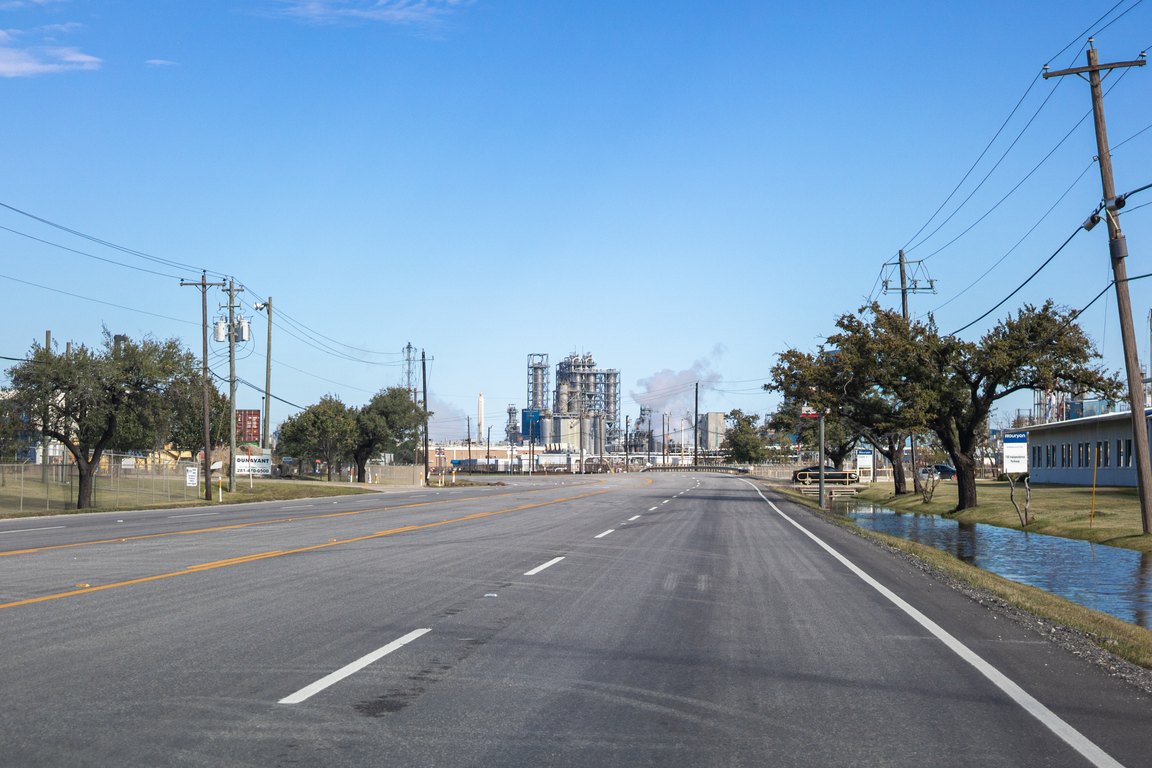 A four-lane road with power lines runs straight toward industrial plant towers under a blue sky.