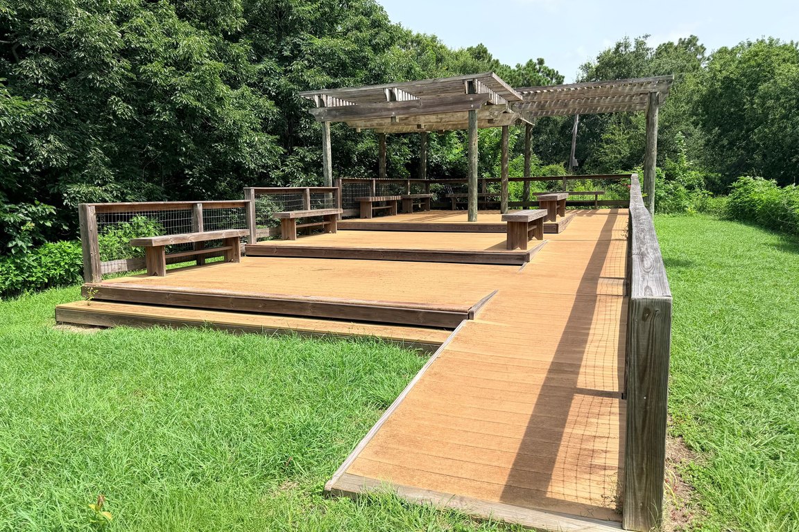 A wooden boardwalk leads to a deck with benches and a pergola, surrounded by grass and dense green trees at Heron Park.