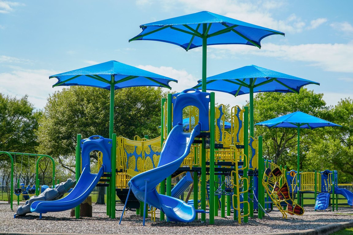 A large playground with blue slides and blue shade umbrellas on a sunny day at Little Cedar Bayou Park.