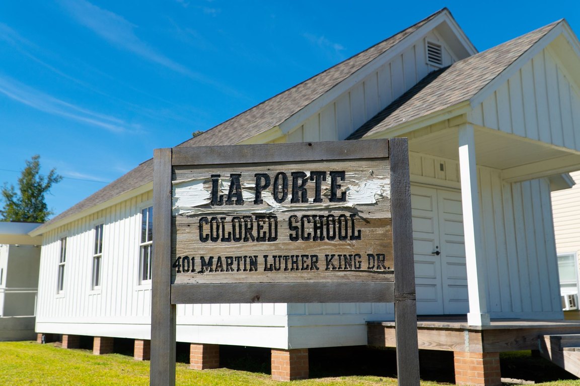 A white one-story wooden schoolhouse with a wooden sign reading La Porte Colored School, 401 Martin Luther King Dr., under a blue sky.