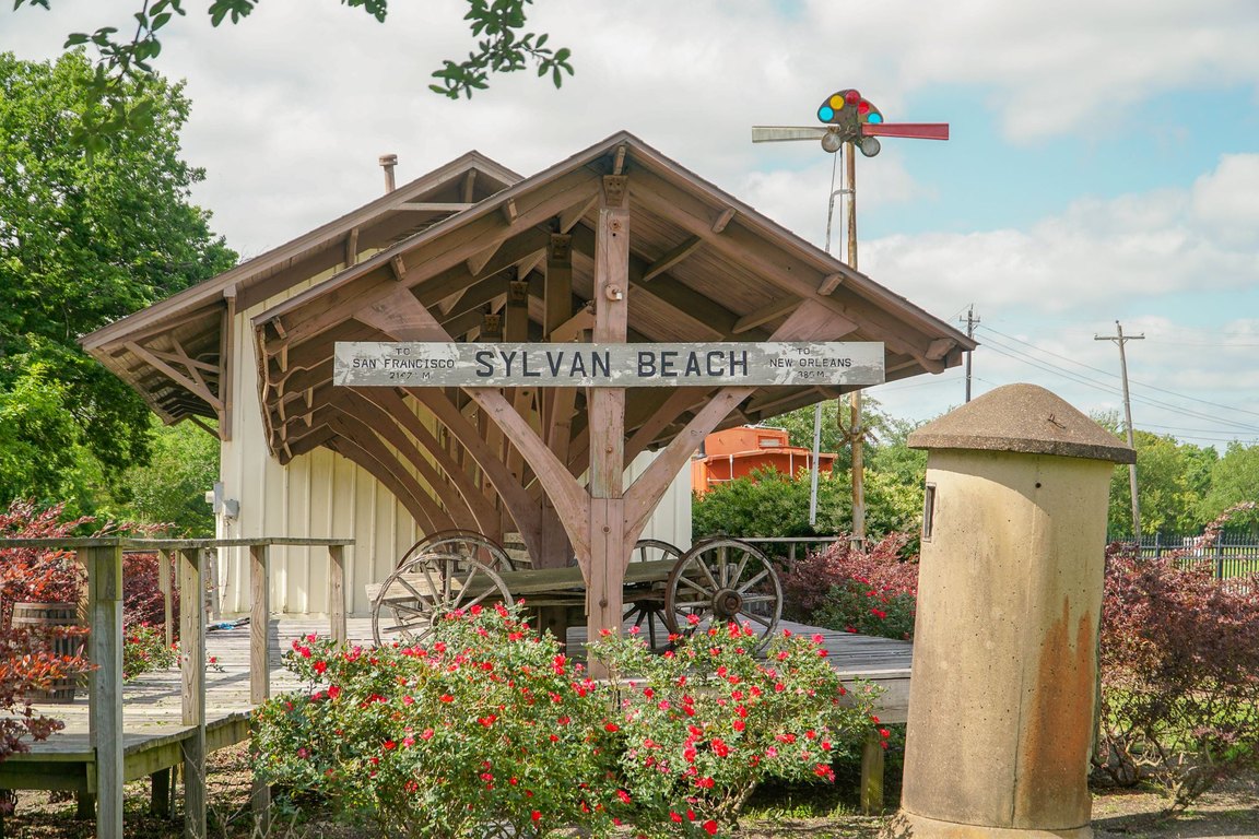A small wooden railroad depot with a Sylvan Beach sign, flowering shrubs and old wagon wheels in front, at Heritage Park.