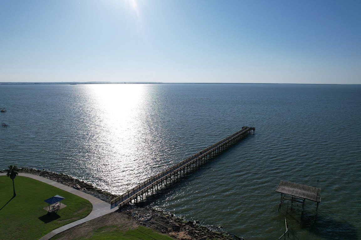 An aerial view of a long fishing pier reaching into sunlit Galveston Bay from the grassy shoreline of Seabreeze Park.