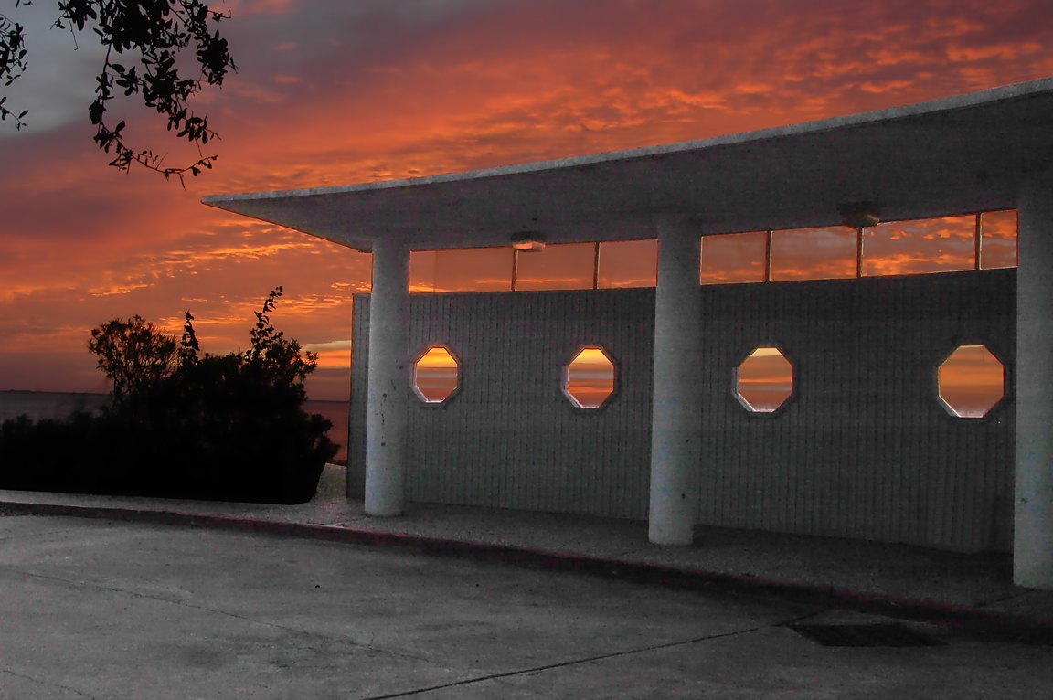 The covered walkway of Sylvan Beach Pavilion, with round porthole windows, silhouetted against an orange sunrise sky over the bay.