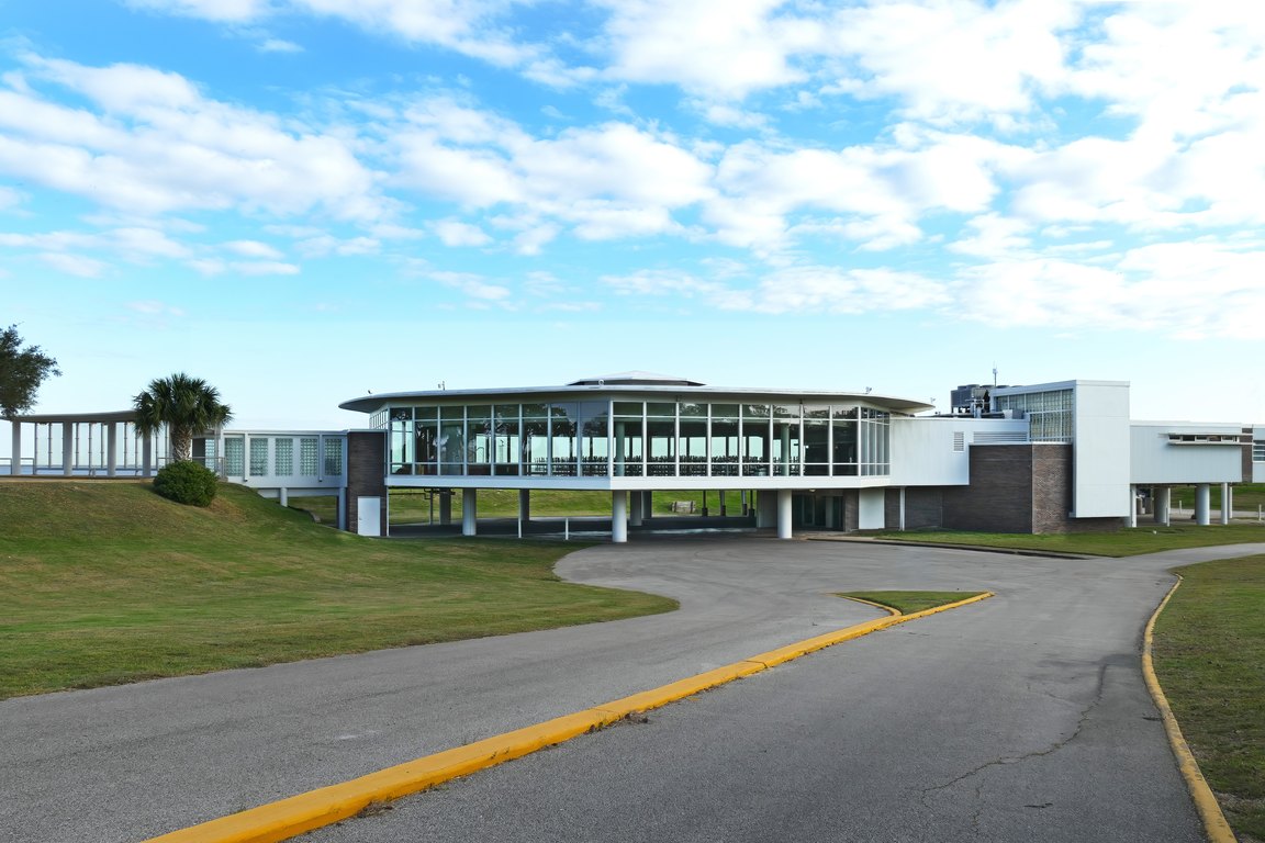 The white mid-century Sylvan Beach Pavilion with a curved glass upper floor, seen from its entry drive under a partly cloudy sky.