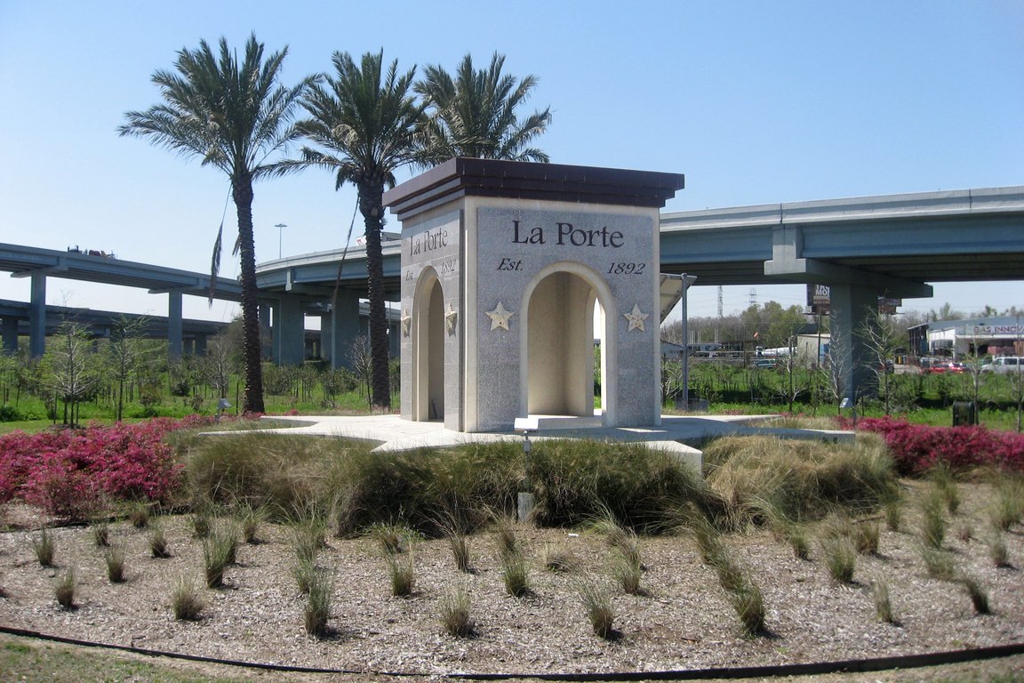 A stone archway monument reading La Porte stands in a landscaped bed with palm trees, with elevated highway ramps behind it.