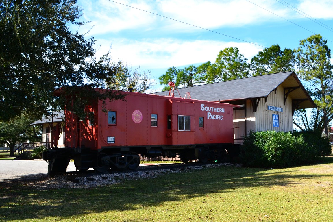 A red Southern Pacific caboose sits on a short track beside a small wooden depot with a Pearland sign, under trees and a blue sky.