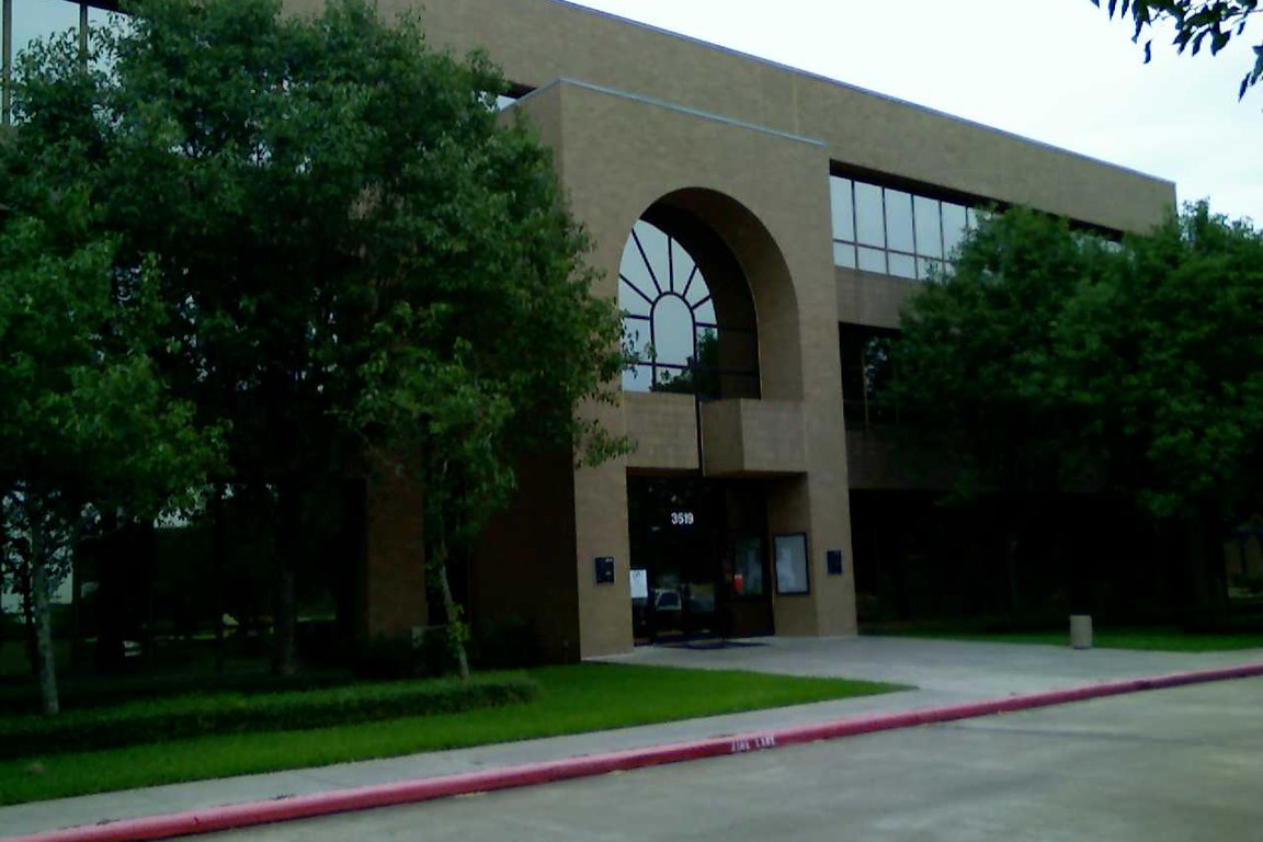 A tan brick office building with a tall arched window over its entrance, framed by trees and a lawn.