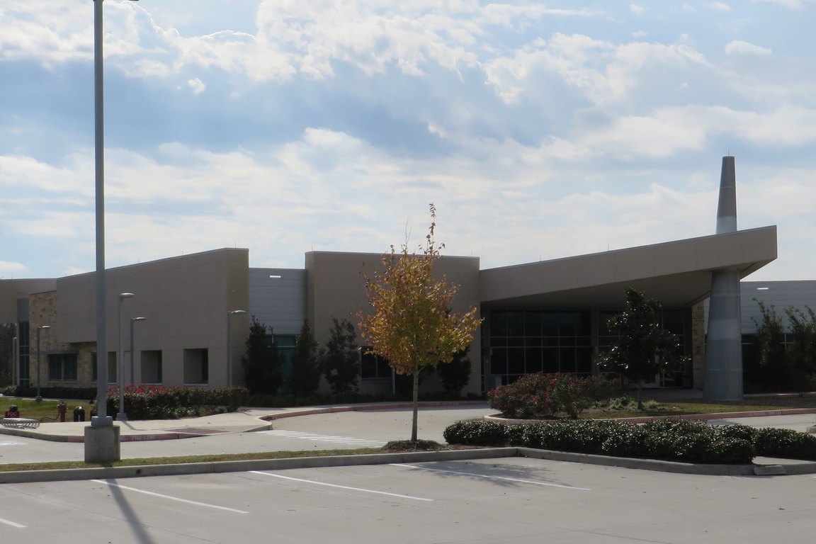 A low modern campus building with a gray canopy entrance and a small tree with autumn leaves beside an empty parking lot.