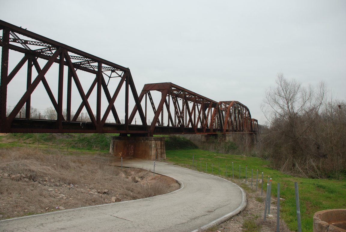 A rust-colored steel truss railway bridge crossing the Brazos River, with a paved path curving beneath it.