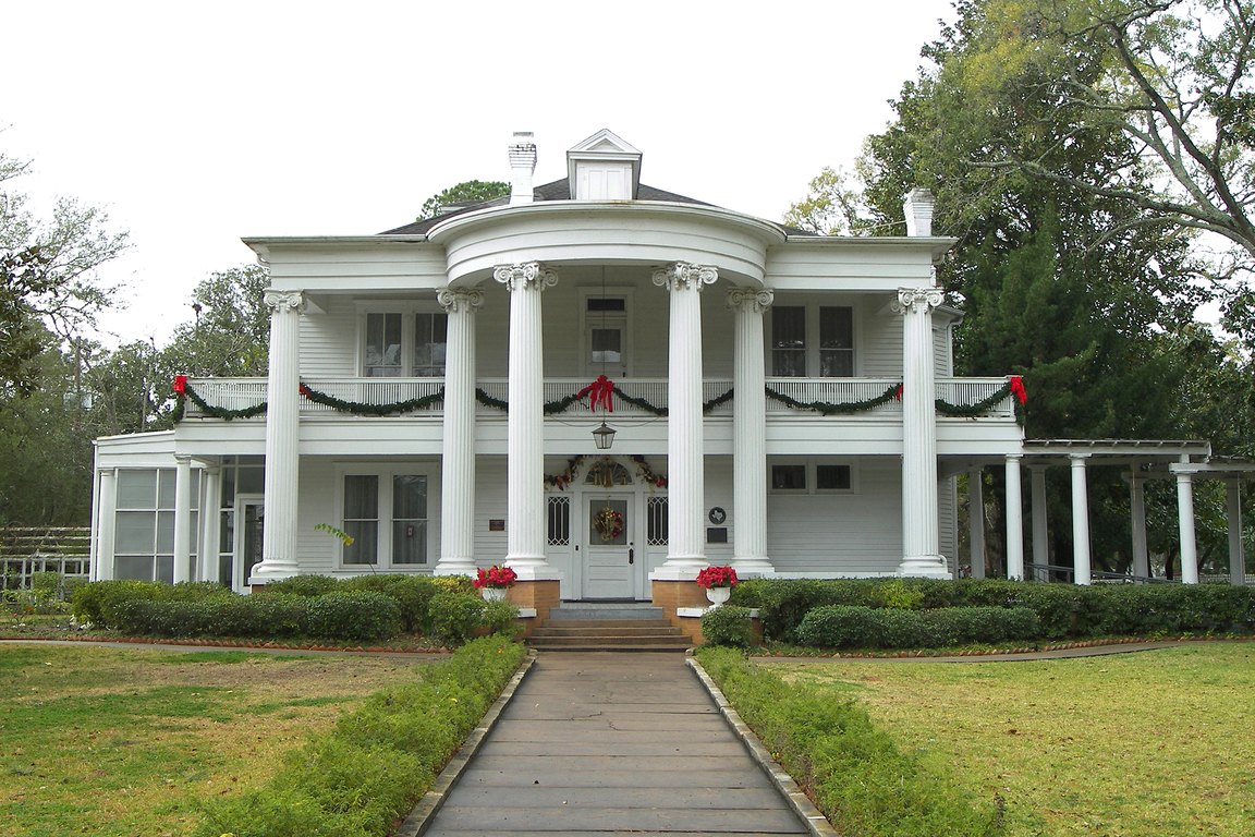 A white two-story house with tall columns and holiday garlands, set behind a lawn and a front walkway.