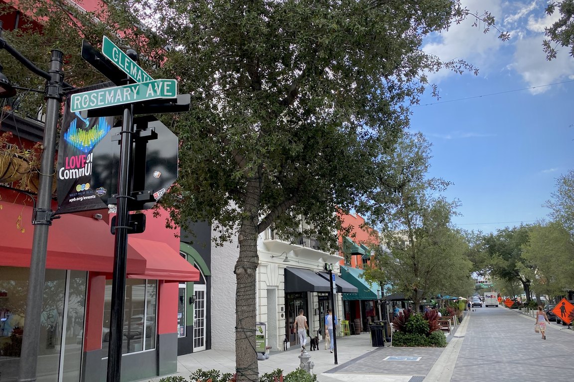 Street signs for Clematis Street and Rosemary Avenue over a tree-lined downtown sidewalk with shops.