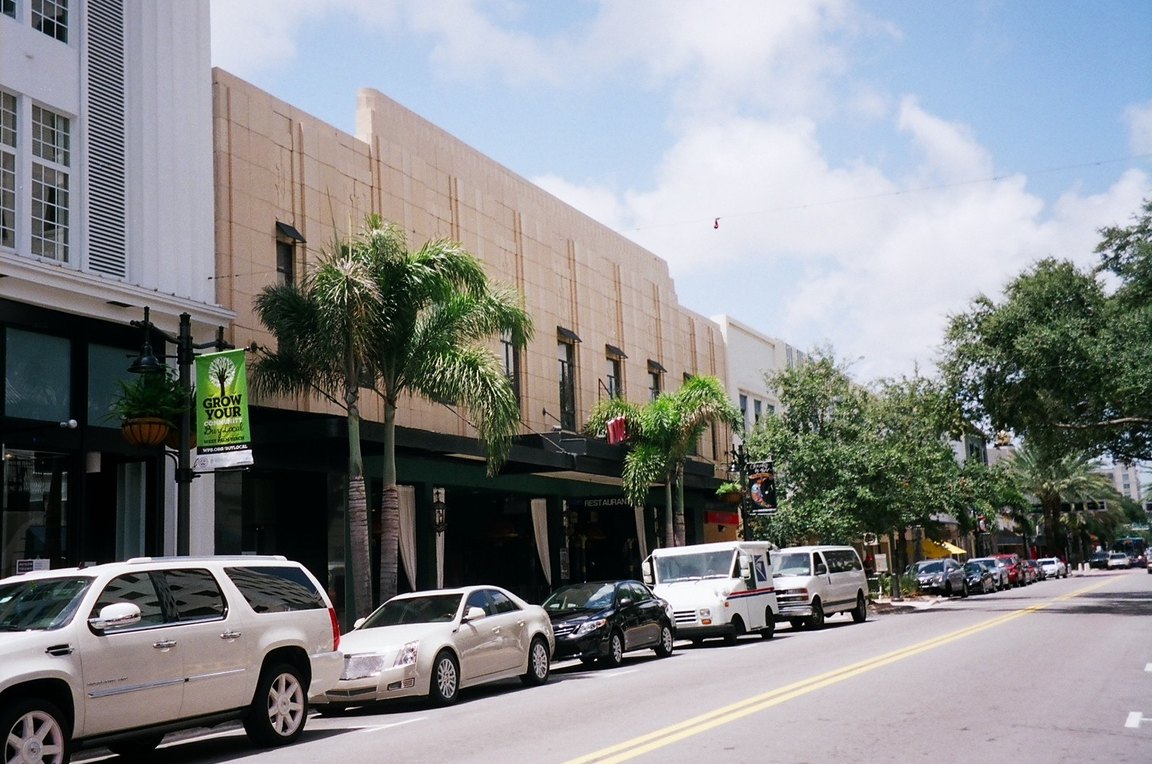 Parked cars along Clematis Street in front of two-story downtown storefronts and palm trees.