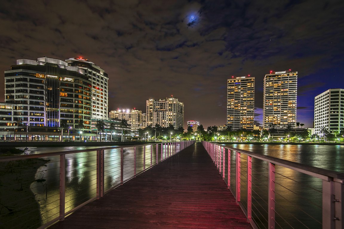 A lit pier over the Intracoastal Waterway at night with downtown West Palm Beach buildings behind it.