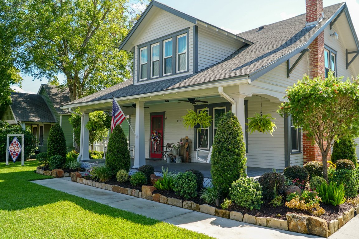 The retreat house for And Sew It Began, a craftsman-style home with a front porch, a flag and landscaped beds.