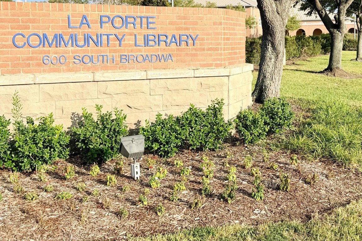 A new landscaping bed with young plants and mulch beside the brick La Porte Community Library sign on South Broadway, under live oak trees.
