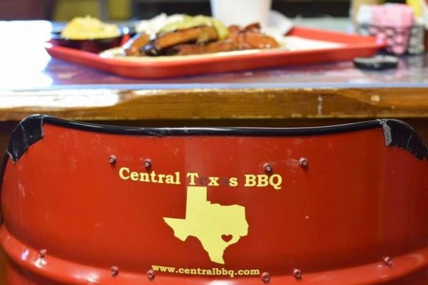 A plate of ribs on a red tray behind a red barrel chair back printed with the Central Texas BBQ name and a Texas outline