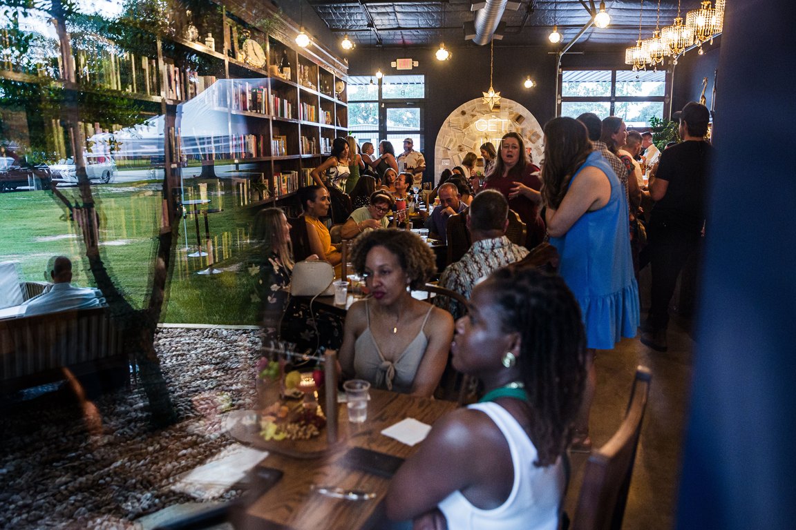 A crowd of neighbors seated and standing inside LIT bookbar, with a wall of bookshelves and a lit arch behind them