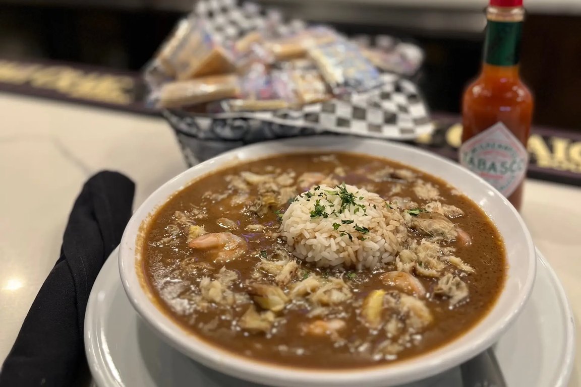 A bowl of gumbo topped with a scoop of white rice on a bar top, with a bottle of hot sauce beside it