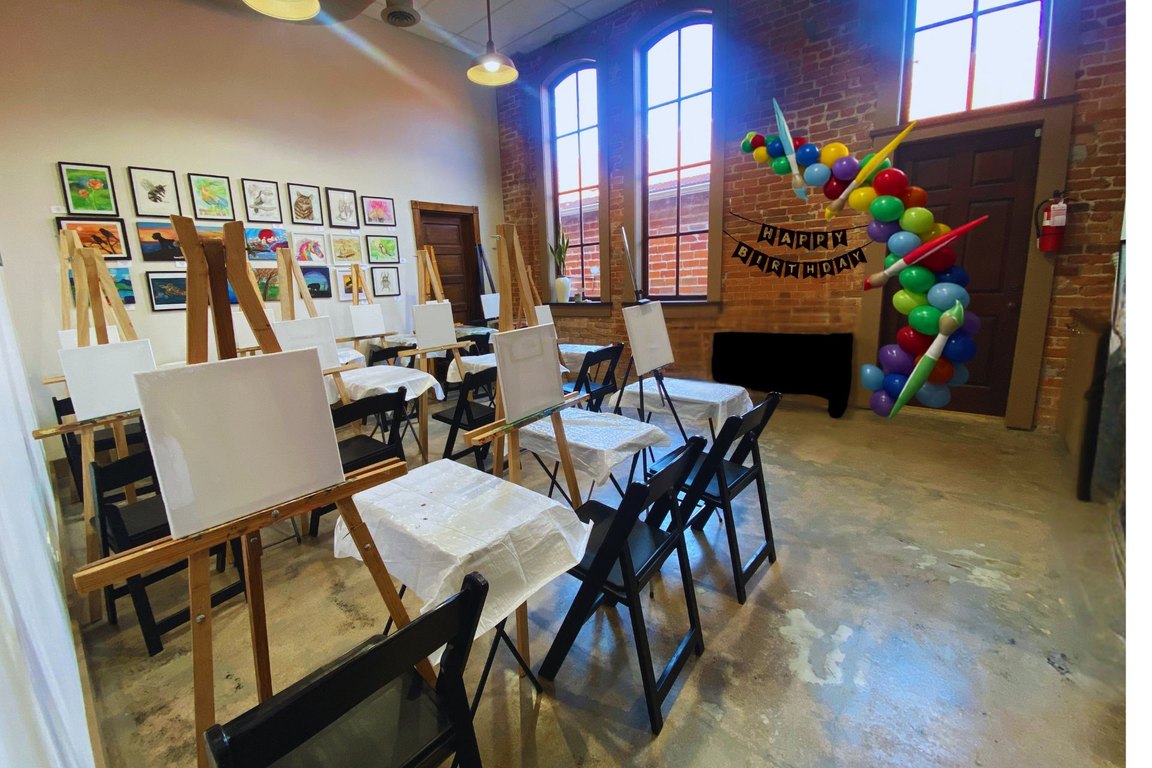 The Catapult Academy art room, with painting easels and tables set up in front of a brick wall with tall windows and a balloon arch