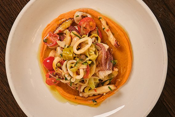 An overhead view of a white plate with sliced squid, cherry tomatoes and herbs over an orange sauce